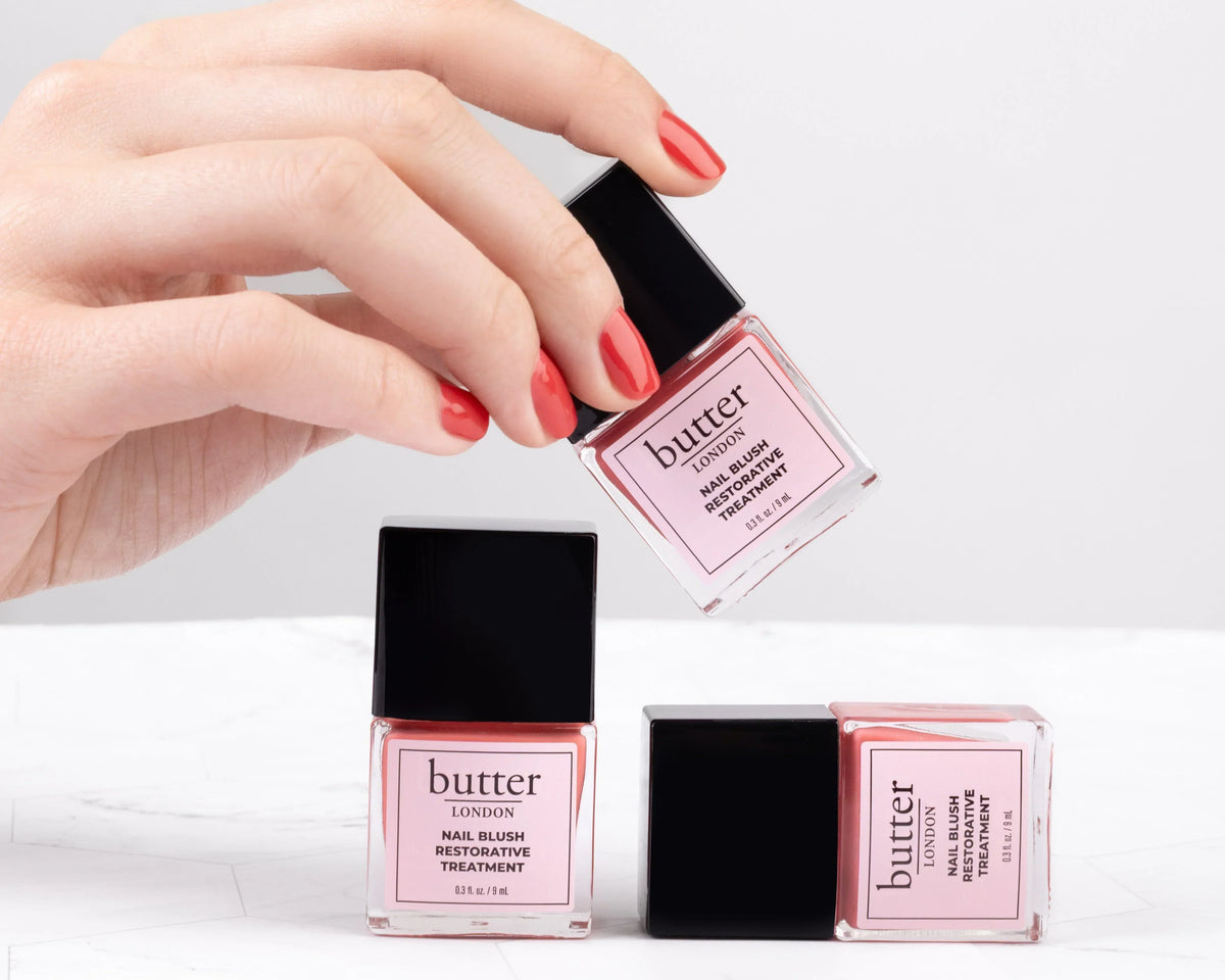 Close up of a hand with coral red nails holding a pink butter LONDON Nail Blush Restorative Treatment bottle