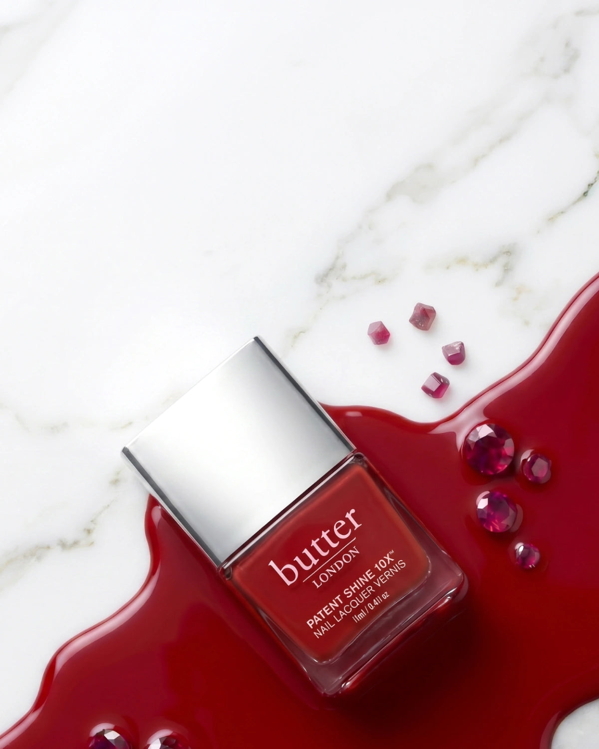 July Colour of the Month - Free Mini Come to Bed Red on $50+: Red PS10X bottle laying in color swatch surrounded by red stones against white marble background (mobile - focal point)